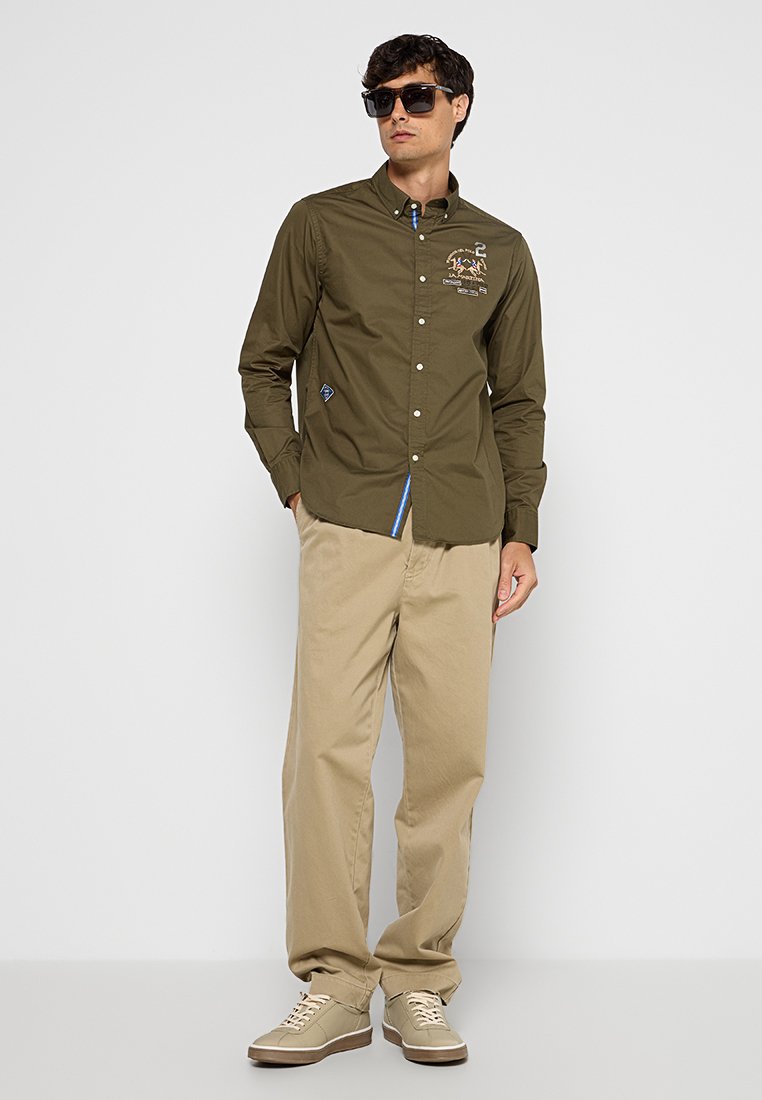 Uomo che indossa una camicia verde scuro con bottoni e un design ricamato, pantaloni beige, sneakers beige e occhiali da sole neri, in piedi contro uno sfondo bianco.