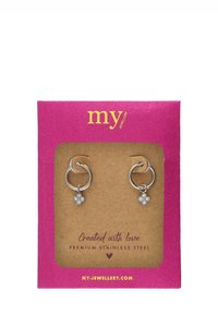 Srebrne kolczyki w formie obręczy ze stali nierdzewnej z wiszącymi kwiatowymi zawieszkami, prezentowane na jasnoróżowej kartce ze złotym tekstem i kraftowym tłem.