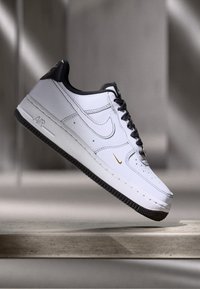 Hvite lær sneakers med svarte detaljer, rund tå og teksturert svart gummisåle. Har et gull Nike swoosh og klassisk snøring.