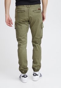 Cargo broeken in olijfgroene katoenmengeling, met twee zijzakken en geribbelde manchetten bij de enkels, gecombineerd met witte sneakers.