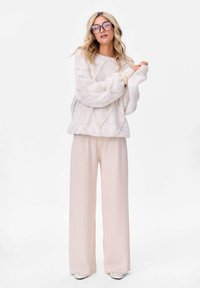 Kobieta z długimi blond włosami, nosząca przesadnie dużą parę okularów, teksturowany kremowy sweter, szerokie beżowe spodnie i jasne buty.