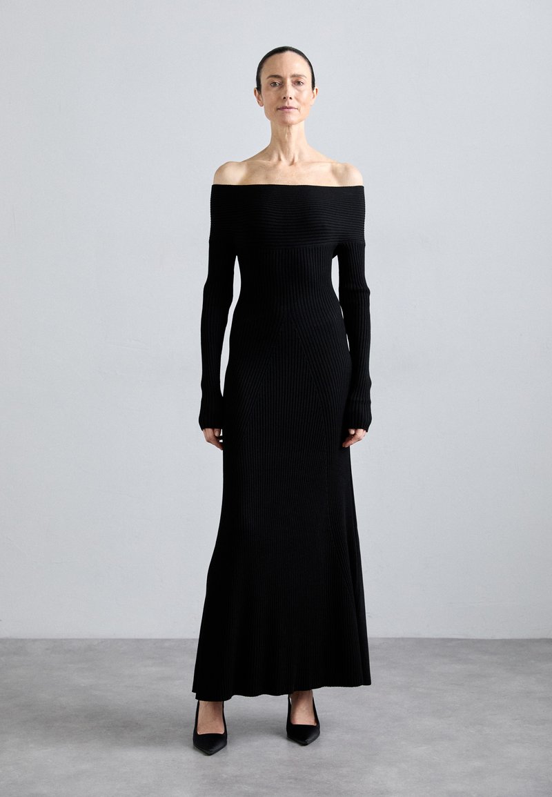 Robe noire côtelée décolletée épaules nues, à manches longues, longueur sol, silhouette près du corps et détails de couture subtils. Chaussures à talons hauts noires.