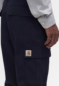 Bărbat purtând pantaloni cargo negri Carhartt și o cămașă gri deschis cu mâneci lungi și manșete cu nasturi, mâna sprijinită pe lângă corp.