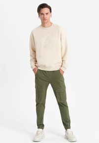 Beige sweatshirt med vit grafisk design, i kombination med olivgröna cargobyxor med fickor. Modellen har på sig ljusa sneakers och står mot en vit bakgrund.