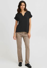 Donna in piedi con una maglietta nera a maniche corte e scollo a V, pantaloni beige a gamba dritta e mocassini neri su uno sfondo chiaro e semplice.