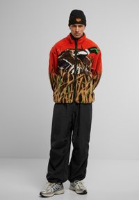 Fleecejakke med en orange overdel med et fugledesign, brun underdel med græs mønstre. Sorte bukser og sølvfarvede sneakers fuldender looket.