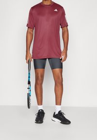 Hombre wearing camiseta atlética color burdeos, pantalones cortos de compresión negros, calcetines blancos y zapatillas negras, sosteniendo una raqueta de tenis negra y azul.