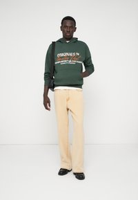 Πράσινη hoodie με άσπρες και πορτοκαλί εκτυπώσεις, κρεμ χρώματος παντελόνι φαρδύ, και μαύρα παπούτσια. Το μοντέλο κρατά μια μικρή μαύρη τσάντα messenger.