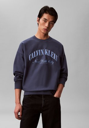Ung mand stående med den ene hånd i lommen på sorte jeans, iført en navyblå Calvin Klein sweatshirt med teksten "New York City" på forsiden.