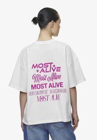 Hvit oversized t-skjorte med rosa grafisk tekst "MOST ALIVE" i ulike skrifttyper og stiler på baksiden. Mykt bomullsmateriale. Avslappet passform.