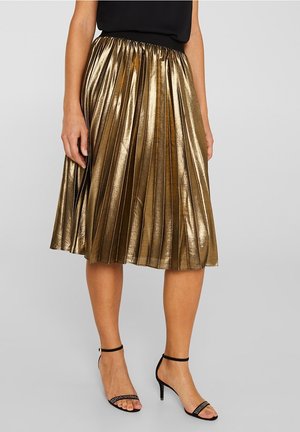Woman wearing a metallic gold pleated midi skirt with a black top and black high-heeled sandals with ankle straps.