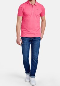 Różowa bawełniana koszulka polo z kołnierzykiem, dwiema guzikami i małym logo; zestawiona z niebieskimi dżinsami i białymi trampkami. Prosty i casualowy design.