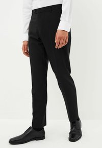 Schwarze maßgeschneiderte Hosen mit glatter Textur, ausgestattet mit einem Knopfverschluss und seitlichen Taschen, kombiniert mit schwarzen, formellen Schuhen.