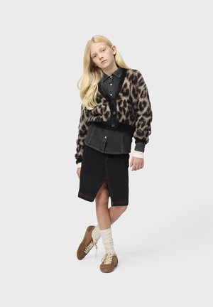 Jeune personne aux longs cheveux blonds portant un cardigan imprimé léopard, une chemise noire boutonnée, une jupe noire fendue à l'avant, des chaussettes crème et des chaussures marron.