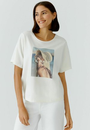 Vrouw draagt een wit T-shirt met een bedrukking van een persoon met een zonneklep bij de zee, glimlachend en staand tegen een effen achtergrond.