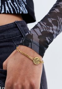 Gold chain bracelet featuring a round charm with intricate detailing, worn on a wrist covered by a long-sleeve mesh top with a black floral pattern.