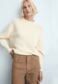 Pull en maille crème avec un col rond et des poignets côtelés, porté avec un pantalon taille haute couleur camel, doté de poches avant et d'un détail avec boutons en métal.