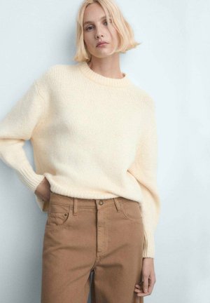 Pull en maille crème avec un col rond et des poignets côtelés, porté avec un pantalon taille haute couleur camel, doté de poches avant et d'un détail avec boutons en métal.