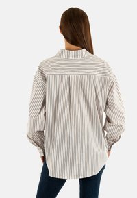 Camisa a rayas en beige claro y marrón, con cuello tipo solapa, mangas largas con volumen y un corte holgado, destacando un detalle de pliegues en la parte posterior.