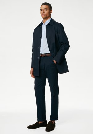 Cappotto trench navy, abbottonato, indossato sopra una camicia azzurro chiaro. Pantaloni navy slim-fit e scarpe in camoscio marrone scuro. Design minimalista, linee pulite.