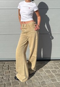 Beige leveleiden corduroy-housut, joissa on korkea vyötärö ja etunapit, kombinert med en kort hvit t-skjorte med en liten rød logo.