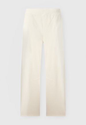 Kobieta o długich ciemnych włosach stoi ze skrzyżowanymi ramionami, ubrana w biały krótki rękaw, kremowe spodnie szerokie na nogawce i beżowe sandały na koturnie.