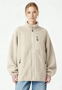 Donna che indossa una giacca in pile beige oversize con tasche sul petto e laterali con cerniera, abbinata a jeans azzurri, in piedi su sfondo bianco.