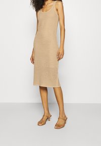 Gebreide midi-jurk in beige met een textuur en open weefpatroon. Voorzien van een ronde halslijn en dunne bandjes. Gecombineerd met transparante hoge hakken.