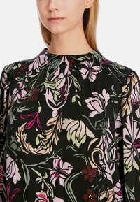 Schwarze Bluse mit floralem Muster, runder Halslinie, langen Ärmeln und einem bunten Muster aus pinken, grünen und burgunderfarbenen Blumen auf dunklem Hintergrund.