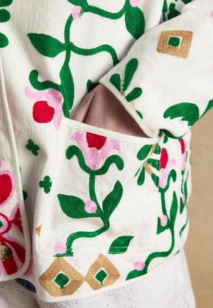 Hand partially in pocket of white fabric with green, pink, red, and gold floral embroidery on jacket sleeve and body.