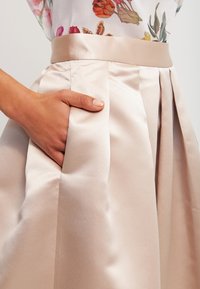 Plisovaná saténová sukně v jemné béžové barvě, s širokým pasem a bočními kapsami, s hladkou, lesklou texturou a elegantním splýváním.