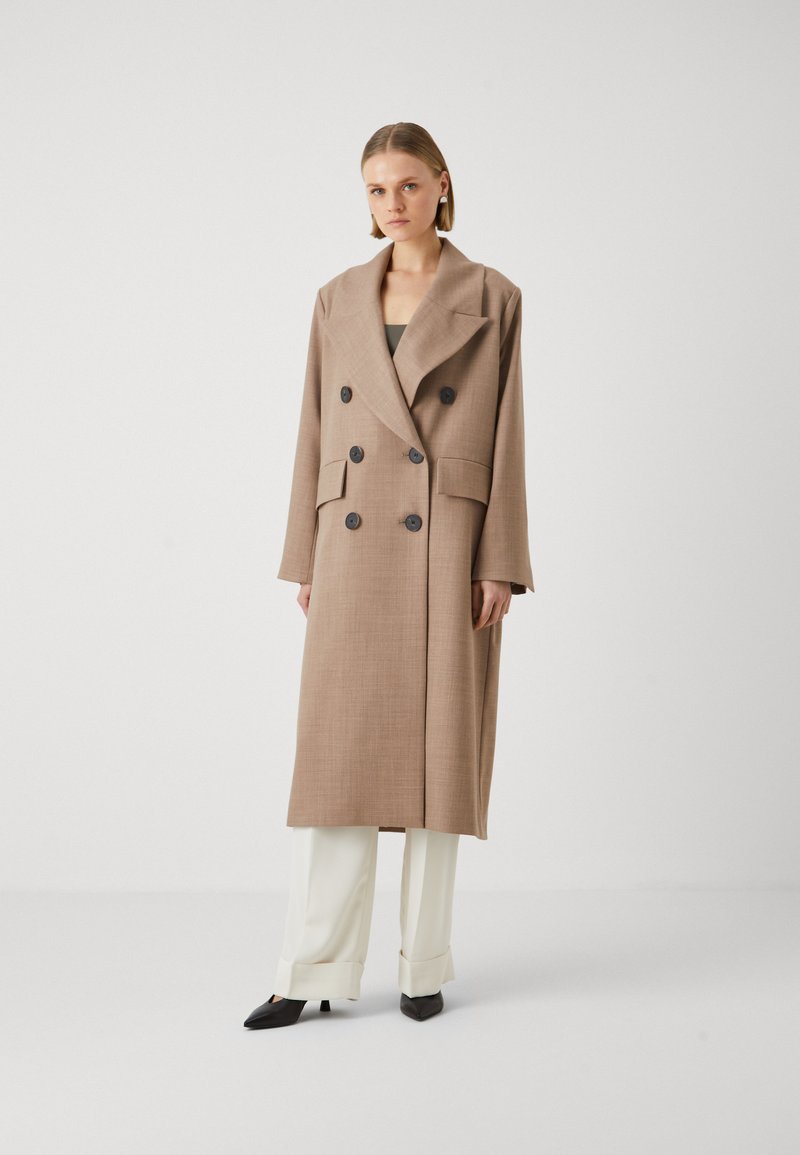 Cappotto lungo doppiopetto marrone realizzato in tessuto testurizzato, con grandi bottoni neri e due tasche frontali. Abbinato a pantaloni bianchi e tacchi neri.