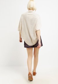 Beżowa, oversize'owa bluzka z drapowanym wzorem i krótkimi rękawami. Figura ma na sobie wzorzystą spódnicę i brązowe buty, widziana z tyłu.