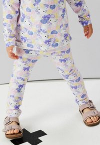 Enfant portant un pyjama pastel à motif Hello Kitty et des sandales roses, debout les jambes écartées sur un sol blanc avec une marque noire en forme de "X".