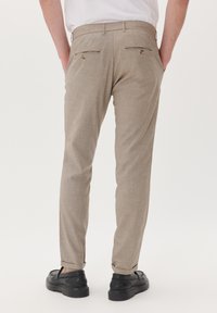 Beige broeken met een gestructureerde afwerking, slim fit ontwerp en opgerolde manchetten, voorzien van twee achterzakken met knoopsluitingen en gedragen met zwarte schoenen.