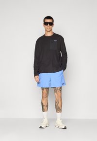 Homme portant une chemise à manches longues noire, un short bleu clair, des chaussettes blanches, des baskets et de grandes lunettes de soleil noires, se tenant devant un fond clair.