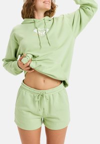 Sudadera de algodón verde claro con un bolsillo frontal y cordón, combinada con pantalones cortos a juego con cordón, que muestran un ajuste relajado.