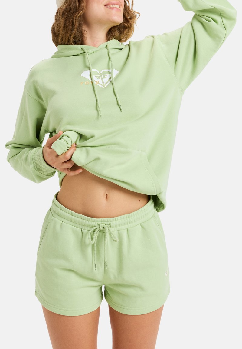 Sudadera de algodón verde claro con un bolsillo frontal y cordón, combinada con pantalones cortos a juego con cordón, que muestran un ajuste relajado.
