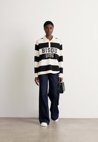 Striped black and white polo shirt with "BISOU 1975" text, paired with wide-leg dark jeans and white shoes, holding a small black handbag.
