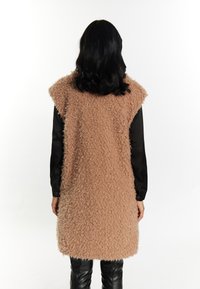 Flauschige beige Weste mit strukturiertem Oberflächen, ärmellosem Design und abgerundetem Saum, kombiniert mit einem schwarzen Langarmshirt und schwarzen Hosen.