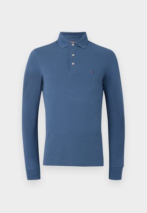 Pitkähihaine sininen polo-paita, jossa on kolme nappia ja pieni logo vasemmassa rinnassa, esitelty valkoista taustaa vasten.