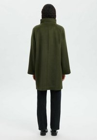 Olijfgroene jas met een hoge kraag, brede mouwen en een recht model. Gecombineerd met donkere jeans en zwarte schoenen. Glad, gestructureerde stof.