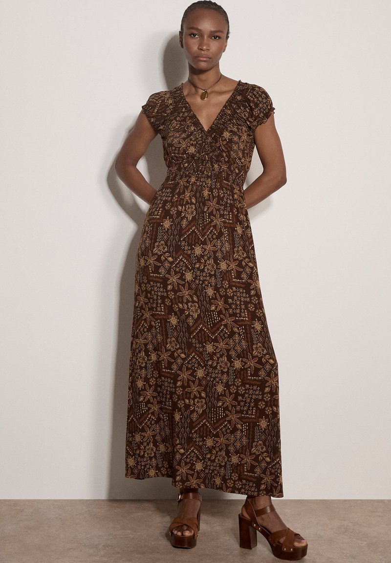 Femme portant une robe longue marron à motif floral, manches courtes et décolleté profond en V, associée à des sandales compensées marron et un collier pendentif en ambre.