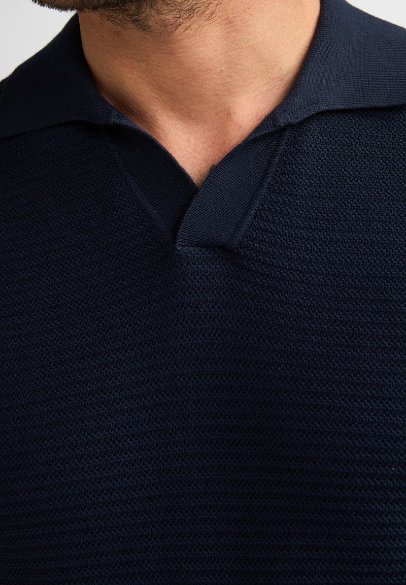 Gros plan sur le cou et la mâchoire d'un homme portant un pull en maille texturée bleu marine foncé avec un large col en V.