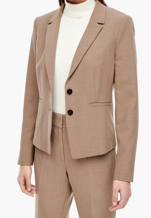 Femme portant un blazer beige sur mesure avec deux boutons noirs et un pantalon taille haute assorti par-dessus un top à col roulé blanc.