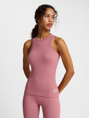 Woman with curly hair in a pink sleeveless ribbed top and matching leggings, standing with hands behind her back, looking to the side.