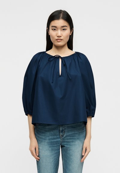 Γυναίκα που φοράει μια φαρδιά μπλε μπλούζα με δέσιμο μπροστά και φουσκωτά μανίκια, συνδυασμένη με ανοιχτόχρωμο τζιν, στέκεται μπροστά σε απλό φόντο.