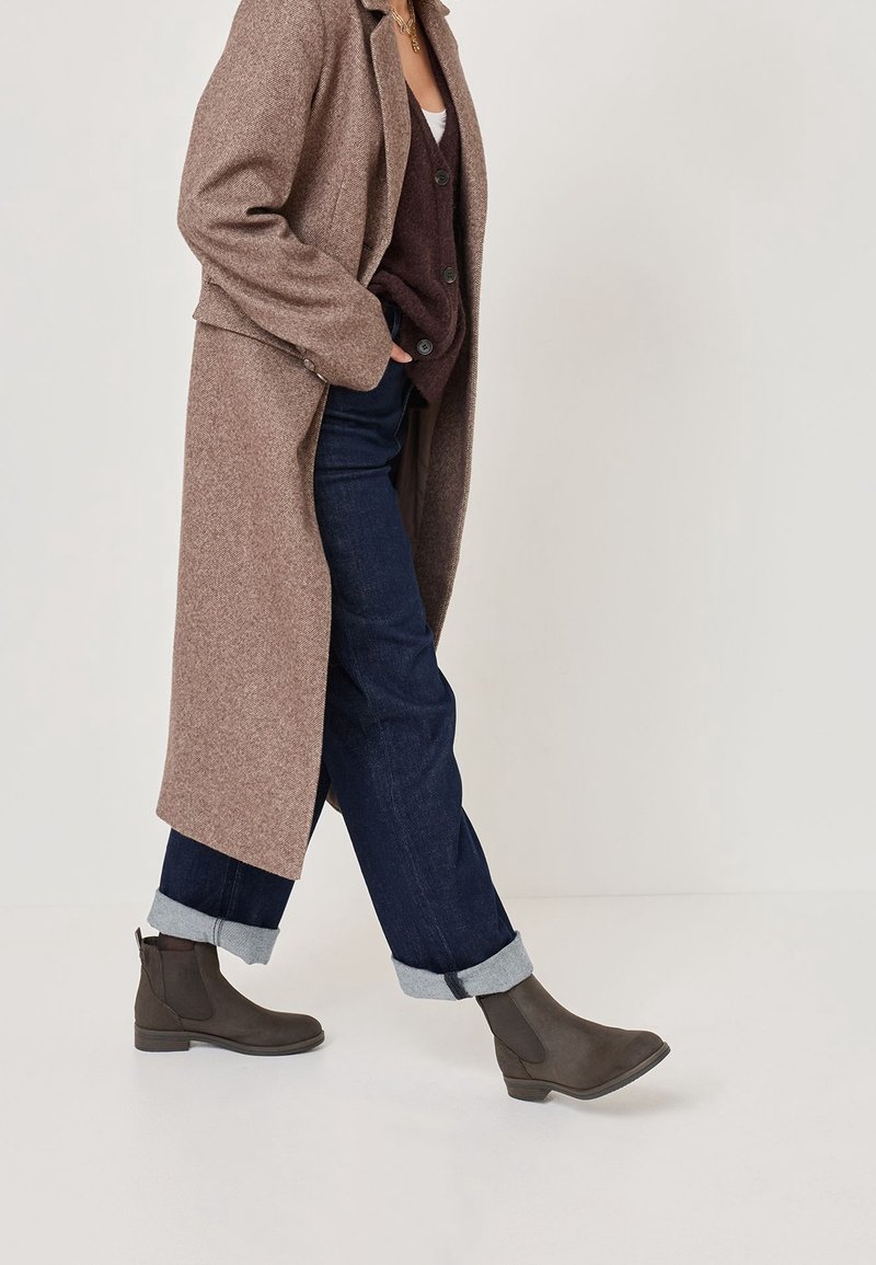 Personne marchant portant un long manteau marron, un gilet sombre boutonné, un jean bleu foncé retroussé et des bottines marron foncé.