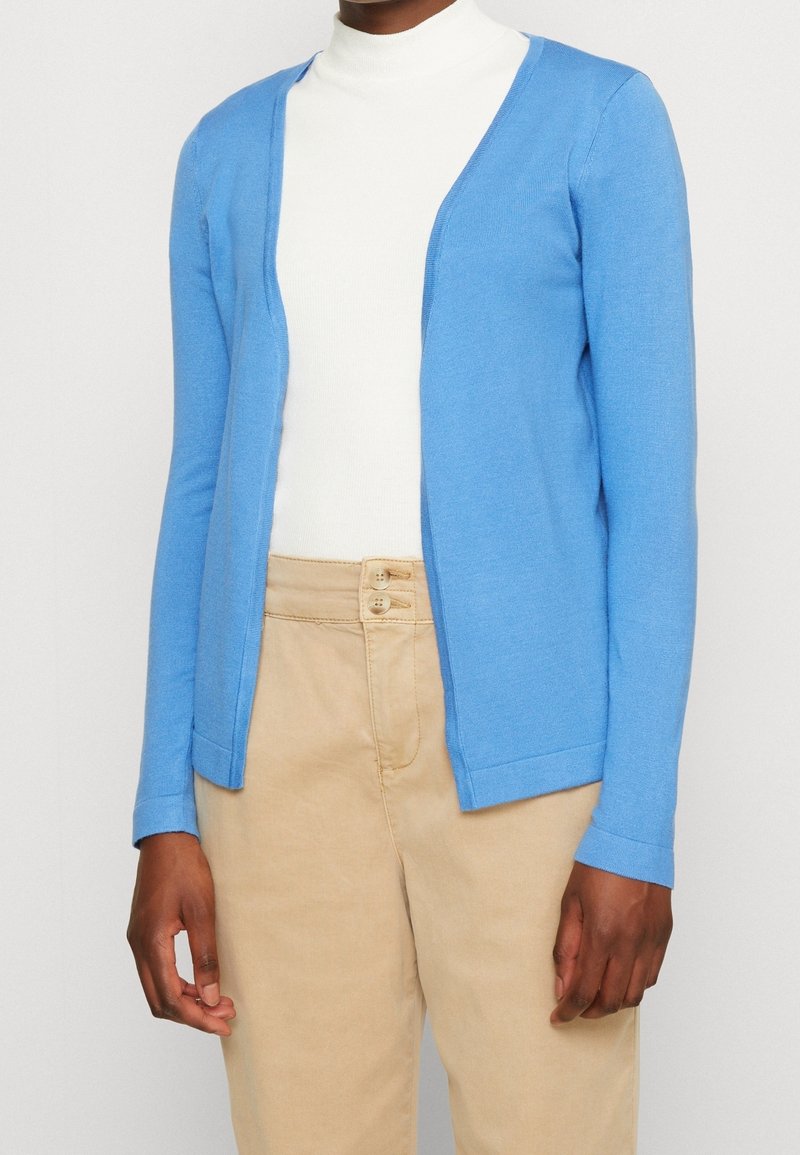 Ljust blå stickad cardigan med öppen front, långa ärmar och ribbade manschetter, bärs över en vit polokrage och beige byxor.