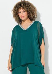 Tüpfelblaues Blusenshirt mit V-Ausschnitt und transparenter, fließender Stoffqualität. Verzierter Ausschnitt mit hellen Perlen. Locker geschnitten mit kurzen Ärmeln.